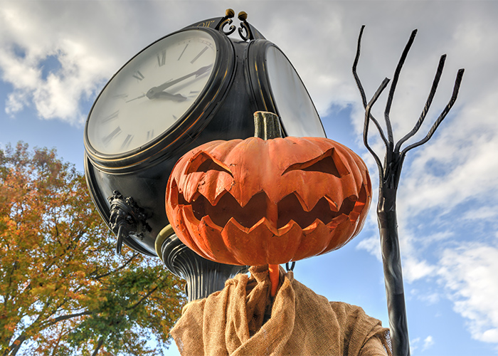 a statue of a pumpkin raising its arm, showing its skinny, stick fingers with a street clock behind it.