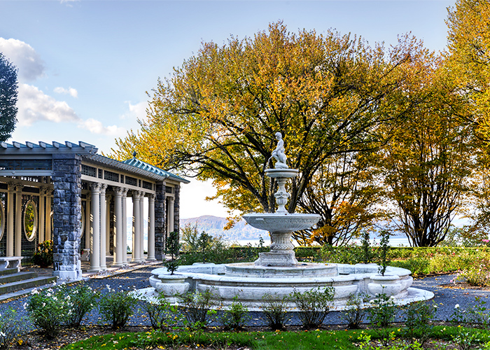 a white fountain with a building to its left with several pillars out front, with the autumn foliage in the background.