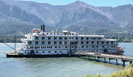 A paddlewheeler cruise ship docked at a port with large mountains in the background.