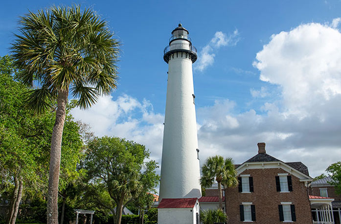 A white lighthouse and a palm tree stand beside each other under a cloudy sky at St. Simons Island Lighthouse Museum. There is a brick building next to the lighthouse and several trees beside it.