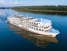 The American Riverboat American Serenade glinding along the Mississippi River, with the tree-filled shore and a bridge in the background. 